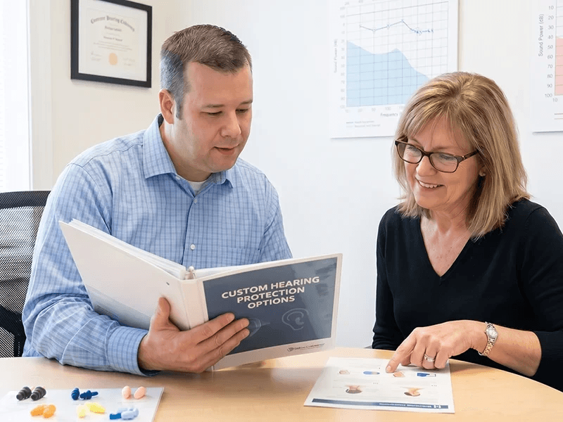 Ryan Holden, Hearing Aid Dispenser, reviewing a custom hearing protection options binder with a client, with sample earplugs and an audiogram visible nearby